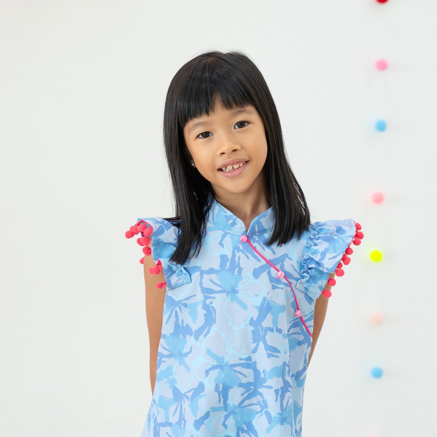 A young girl wearing a light blue ruffle cheongsam dress with pink pom-pom trim from the Le Petit Society x Sheila Sim CNY 2026 collection smiles softly at the camera. Colourful hanging pom-poms decorate the bright background.