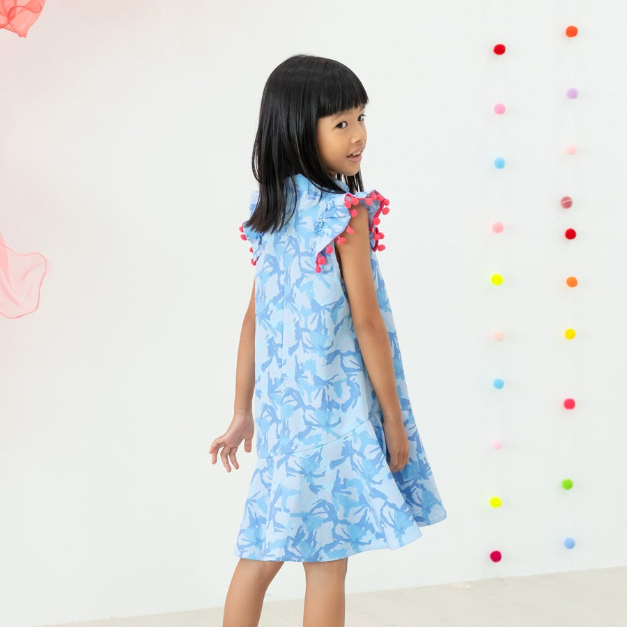 A young girl wearing a light blue ruffle cheongsam dress with pink pom-pom trim from the Le Petit Society x Sheila Sim CNY 2026 collection turns slightly to look back over her shoulder. She stands against a bright backdrop decorated with colourful hanging