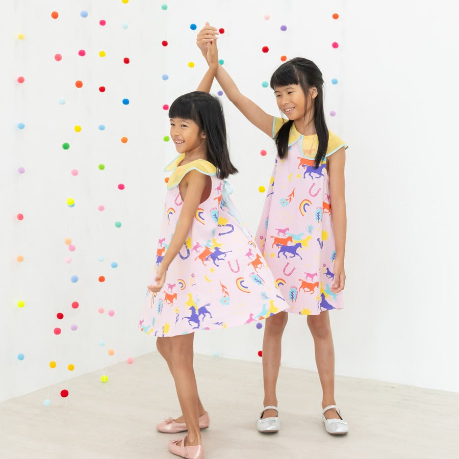 Two girls dance and twirl in their Rainbow Horse dresses, holding hands with bright smiles. The A-line silhouettes flare softly as they move, showcasing the vibrant horse, rainbow and star illustrations. The colourful hanging pom-poms create a joyful, cel