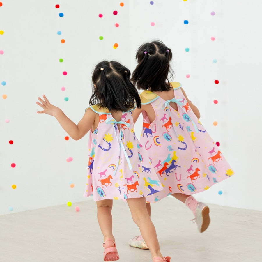 A back view of two girls playfully running together in their Rainbow Horse dresses. The dresses feature sweet ribbon ties and scalloped collars, with the colourful horse and lucky-charm motifs swaying as they move. The trailing motion of the girls and the