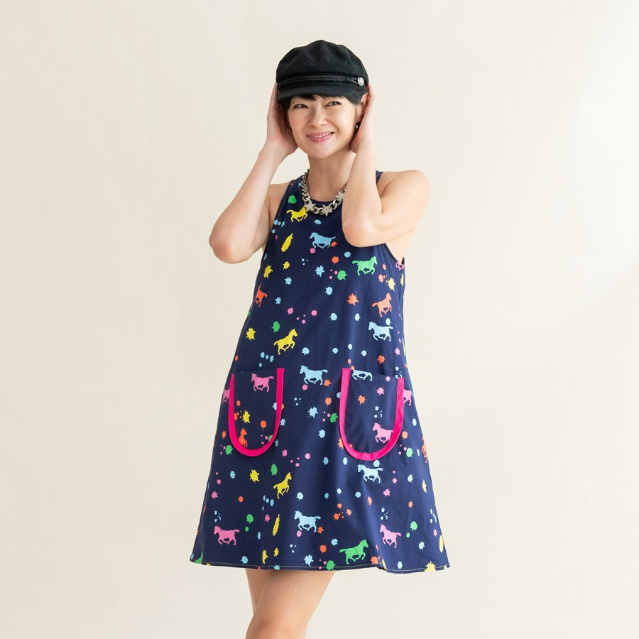A woman adjusting her black cap while wearing the sleeveless navy dress from the Le Petit Society x Sheila Sim CNY 2026 Rainbow Horse Collection, featuring bright rainbow horse and splatter prints.