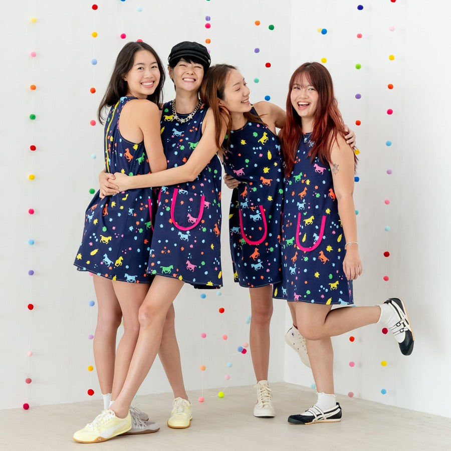 Four women in matching navy dresses from the Le Petit Society x Sheila Sim CNY 2026 Rainbow Horse Collection, hugging and laughing together in front of a wall decorated with colourful pom-pom strings.
