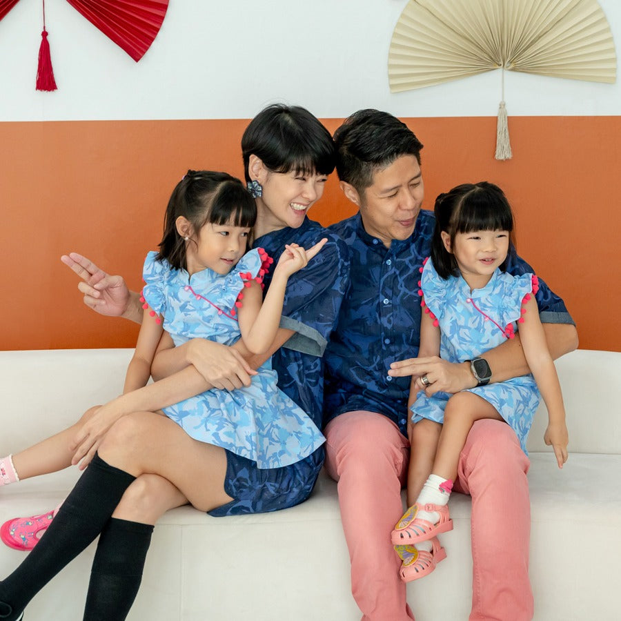 A family of four wearing matching outfits from the Le Petit Society x Sheila Sim CNY 2026 collection sits together on a white sofa. The parents wear navy blue chambray pieces while the two young girls wear light blue ruffle cheongsam dresses with pink pom