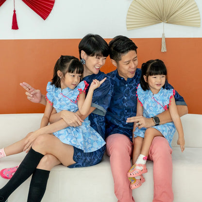 A family of four wearing matching outfits from the Le Petit Society x Sheila Sim CNY 2026 collection sits together on a white sofa. The parents wear navy blue chambray pieces while the two young girls wear light blue ruffle cheongsam dresses with pink pom