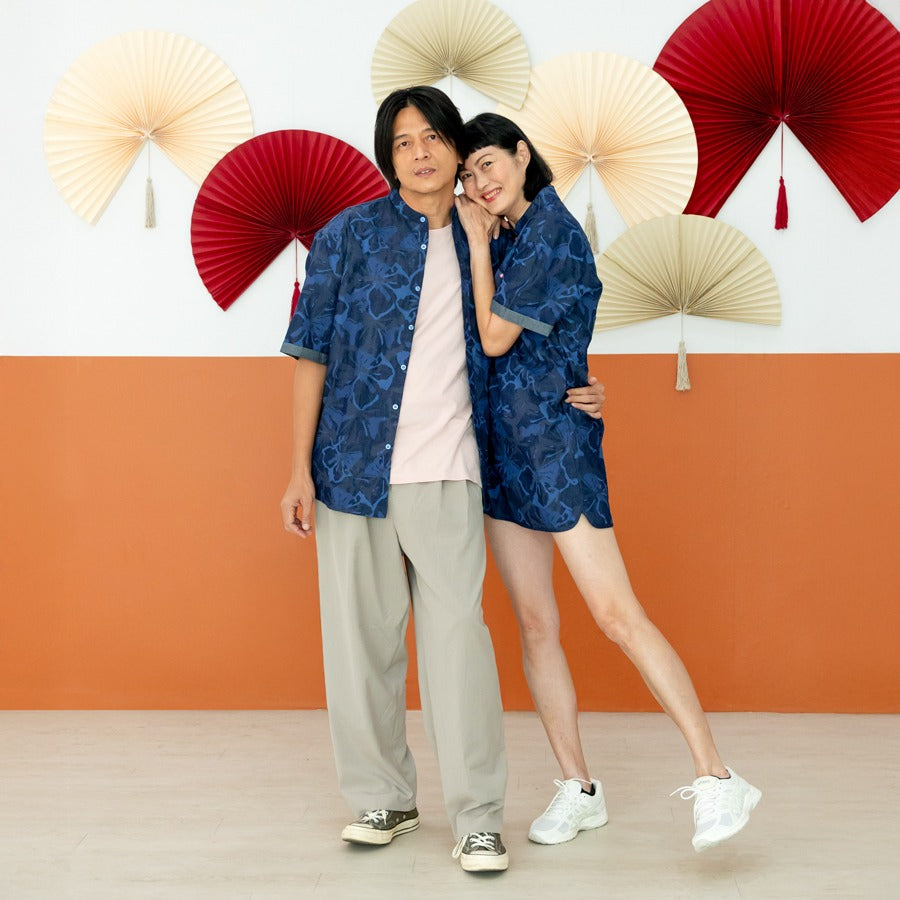 A male model in a navy blue chambray floral short-sleeve shirt and a female model in a matching chambray floral cheongsam-inspired dress from the Le Petit Society x Sheila Sim CNY 2026 collection stand together, with the female model leaning affectionatel