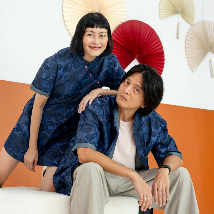 A female model in a navy blue chambray floral cheongsam-inspired dress and a male model in a matching chambray shirt from the Le Petit Society x Sheila Sim CNY 2026 collection pose together on a white sofa. The female model leans forward with her hand on 