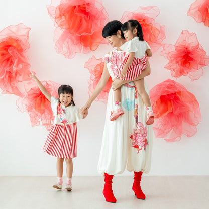 A mother and her two young daughters wear matching pieces from the Le Petit Society x Sheila Sim CNY 2026 lotus collection. The mother stands in a cream jumpsuit with a large floral print, holding one daughter while the other raises her arm excitedly. Bot