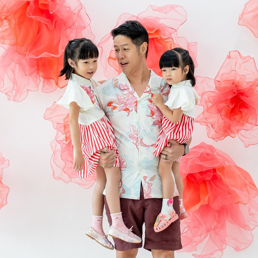 A dad carries his two daughters, all dressed in matching pieces from the Le Petit Society x Sheila Sim CNY 2026 Lotus Collection. He wears the men’s lotus-print shirt in soft coral and blue tones, while the girls are in the coordinating lotus-and-stripe c