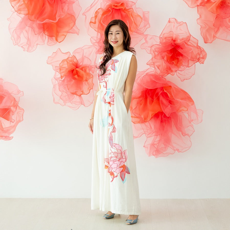 A female model wearing a long cream dress with large pink and blue lotus floral prints from the Le Petit Society x Sheila Sim CNY 2026 collection stands smiling against a white wall decorated with soft coral organza flower accents. She completes the look 