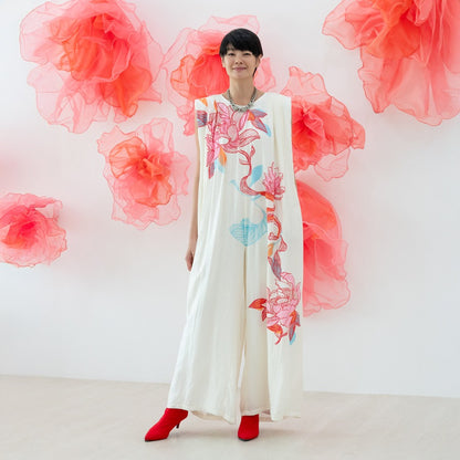 A female model wearing a cream lotus-print jumpsuit with wide, gathered legs from the Le Petit Society x Sheila Sim CNY 2026 collection stands smiling against a white wall decorated with large coral organza flowers. She pairs the look with bold red heeled