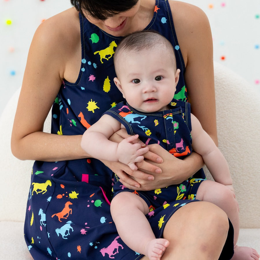 Le Petit Society x Sheila Sim CNY 2026 Rainbow Horse Collection — A baby sits comfortably on an adult’s lap, both wearing matching navy outfits covered in colourful rainbow horse prints. The baby looks toward the camera while soft pom-pom decor adds a che