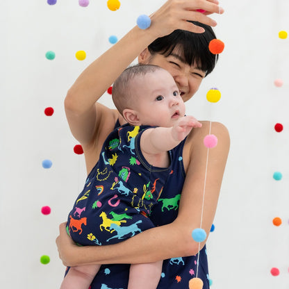 Le Petit Society x Sheila Sim CNY 2026 Rainbow Horse Collection — A baby in a navy horse-print romper reaches out eagerly toward a string of colourful pom-poms while being held. The playful backdrop of floating pom-poms adds a whimsical, festive feel.