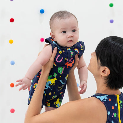Le Petit Society x Sheila Sim CNY 2026 Rainbow Horse Collection — A baby wearing a sleeveless navy romper decorated with colourful rainbow horses and lucky icons is lifted gently in the air. The baby looks wide-eyed and curious, with strands of bright pom