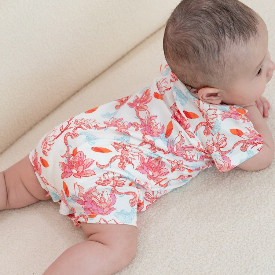 A baby lies comfortably on a soft cream surface, wearing the lotus-print romper from the Le Petit Society x Sheila Sim CNY 2026 collection. The back view shows off the full lotus artwork in flowing corals, pinks, and touches of blue — a gentle, dreamy pri
