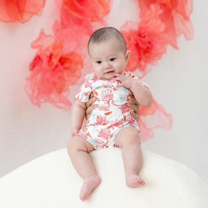 A tiny baby is being gently held up, dressed in a sweet lotus-print romper from the Le Petit Society x Sheila Sim CNY 2026 lotus collection. The floral pattern in soft corals and blues looks super delicate on the little one, and the flutter sleeves give i
