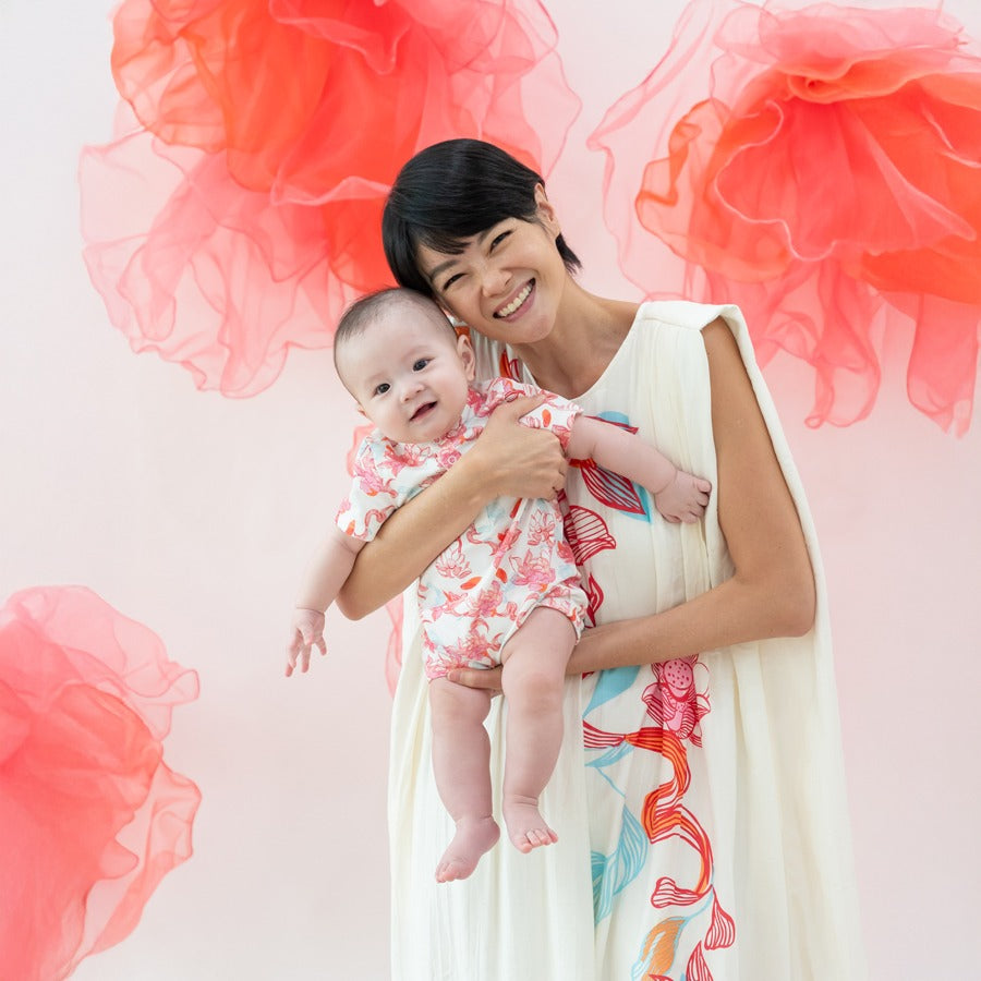 A mum beams as she carries her little one, both dressed in matching pieces from the Le Petit Society x Sheila Sim CNY 2026 lotus collection. The baby’s lotus-print romper looks extra sweet against the airy cream dress worn by Mum, featuring the same flowi