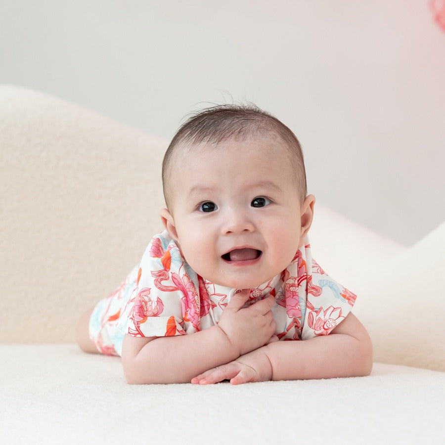 A sweet baby lies tummy-down in a cosy setting, dressed in the lotus-print romper from the Le Petit Society x Sheila Sim CNY 2026 collection. The soft coral and blue floral artwork stands out beautifully against the baby’s bright, curious expression, givi