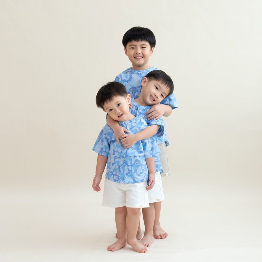 Three young boys wearing matching light blue printed shirts from the Le Petit Society x Sheila Sim CNY 2026 collection stand in a playful stacked hug, leaning gently against one another. The older boy stands behind the two younger ones, all smiling agains