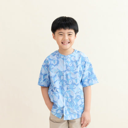 A young boy wearing a light blue printed short-sleeve shirt from the Le Petit Society x Sheila Sim CNY 2026 collection stands smiling with one hand in his pocket against a soft cream background. He is dressed in beige shorts and looks cheerful and relaxed