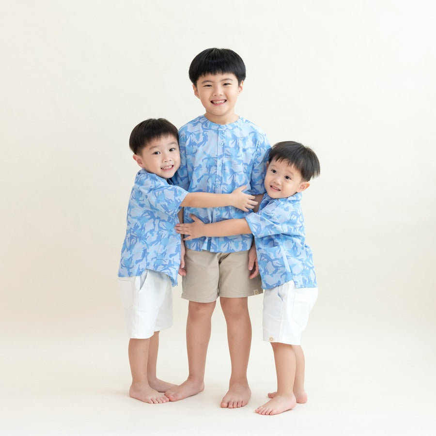 Three young boys wearing matching light blue printed shirts from the Le Petit Society x Sheila Sim CNY 2026 collection stand together in a group hug against a soft cream background. The older boy is in the center wearing beige shorts, while the two younge