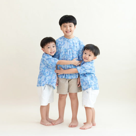 Three young boys wearing matching light blue printed shirts from the Le Petit Society x Sheila Sim CNY 2026 collection stand together in a group hug against a soft cream background. The older boy is in the center wearing beige shorts, while the two younge