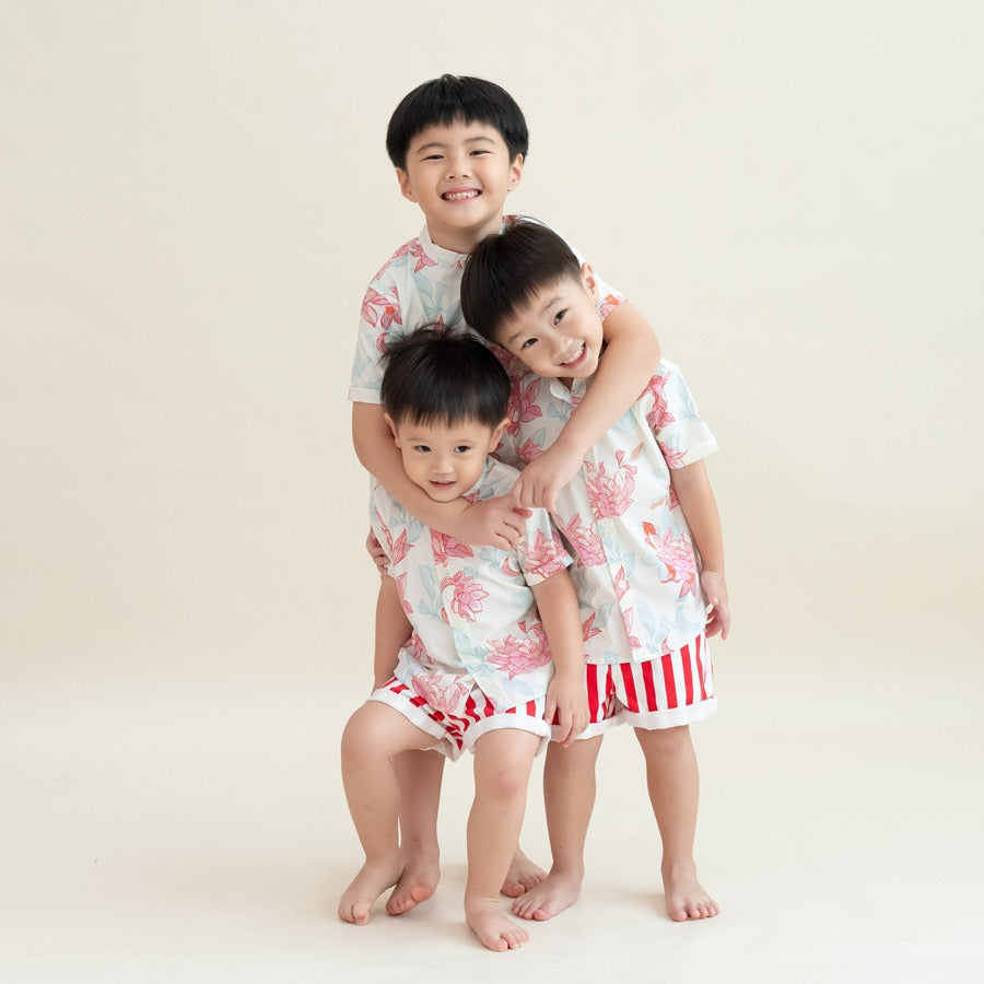 Three boys in the matching Lotus Collection outfits stand close together, hugging and smiling. The coordinated shirts and bold striped shorts create a bright, cheerful palette, while their warm interaction captures the sweetness of family bonding during C