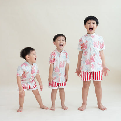 Three boys dressed in matching Lotus Collection outfits stand in a row, bursting into laughter. Their mandarin-collar shirts are covered in coral and pastel lotus illustrations, complemented by bold red-and-white striped shorts. The joyful energy of the m