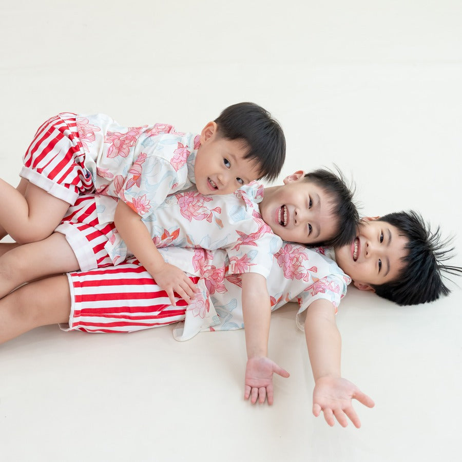 Three boys pile playfully on top of one another while wearing the Lotus Collection shirts and striped shorts. Their joyful expressions and relaxed poses bring out the fun, carefree mood of Lunar New Year celebrations, while the coral and sky-blue lotus mo