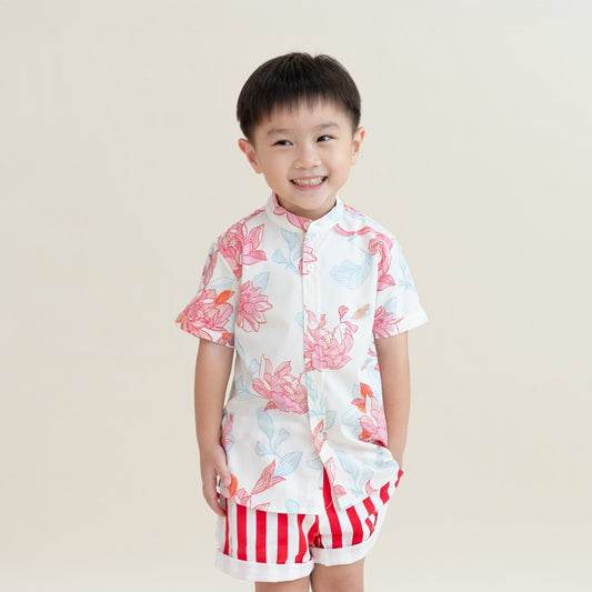 A young boy wears the boys’ shirt and shorts from the Le Petit Society x Sheila Sim CNY 2026 Lotus Collection. The mandarin-collar shirt features coral and sky-blue lotus blooms against a soft white base, paired with playful red-and-white striped shorts. 