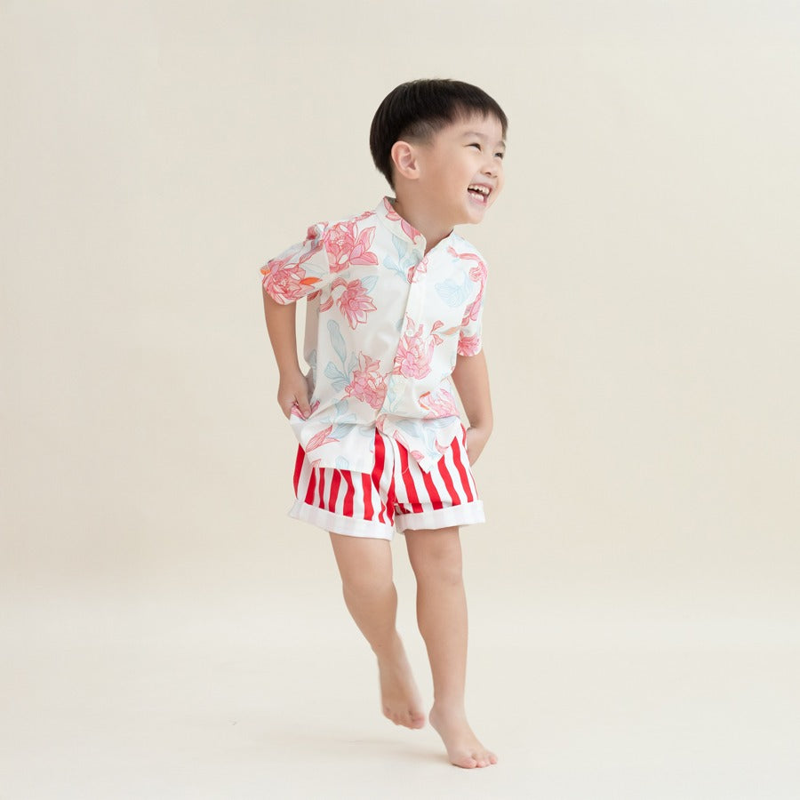 A young boy wearing a white short-sleeve shirt with pink and blue lotus floral prints and red-and-white vertical striped shorts from the Le Petit Society x Sheila Sim CNY 2026 collection runs joyfully against a soft cream background, smiling with his hand