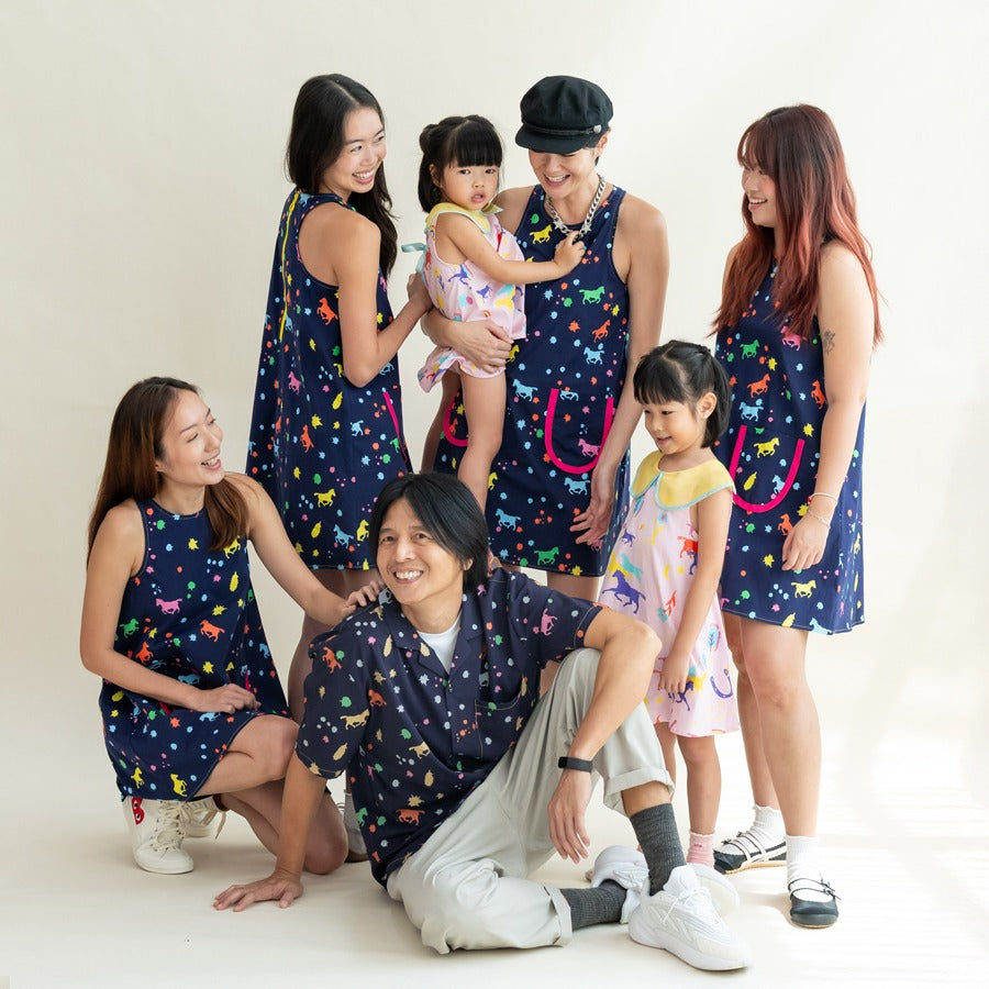 A group of adults and children gather in coordinated Rainbow Horse pieces, smiling and interacting naturally. The women wear the navy dresses, while the children mix navy and pastel pink prints. The relaxed, happy energy shows the playful, family-friendly