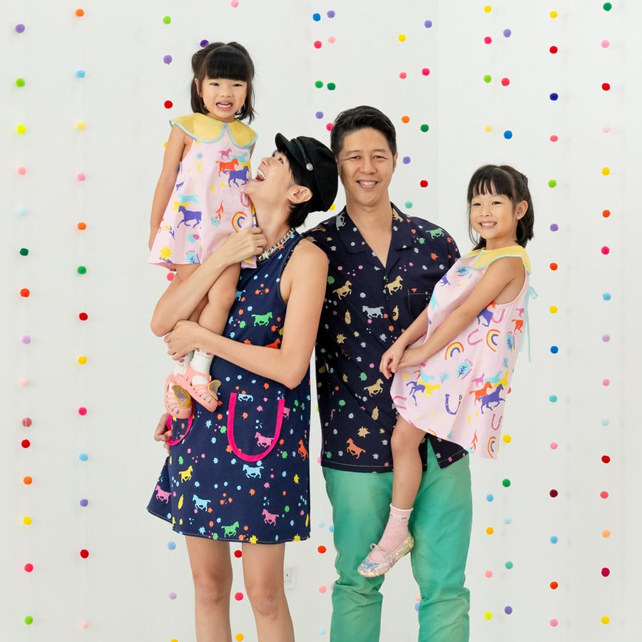 A family of four dresses in the Rainbow Horse pieces from the Le Petit Society x Sheila Sim CNY 2026 Collection. The father wears the navy men’s shirt, the mother the matching navy dress, while the daughters wear pastel pink Rainbow Horse dresses. Bright 