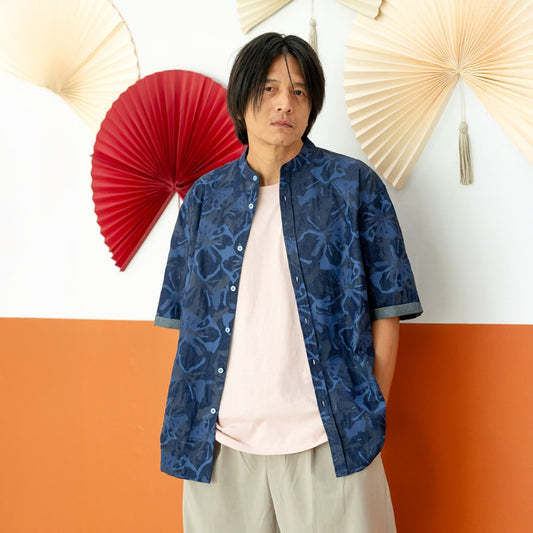 A male model wearing a navy blue chambray floral short-sleeve shirt from the Le Petit Society x Sheila Sim CNY 2026 collection stands with his hands in his pockets in front of a two-tone orange and white wall decorated with red and cream paper fans. He fa