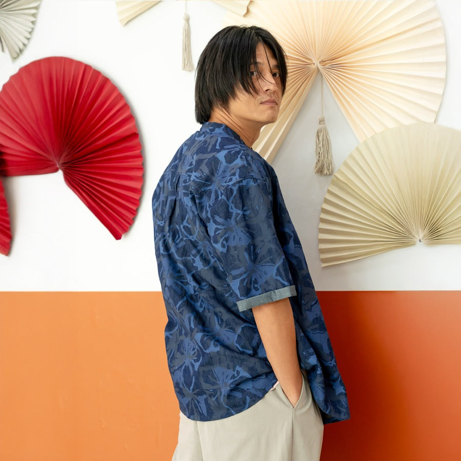 A male model wearing a navy blue chambray floral short-sleeve shirt from the Le Petit Society x Sheila Sim CNY 2026 collection stands turned slightly away from the camera with his hands in his pockets, in front of a two-tone orange and white wall decorate