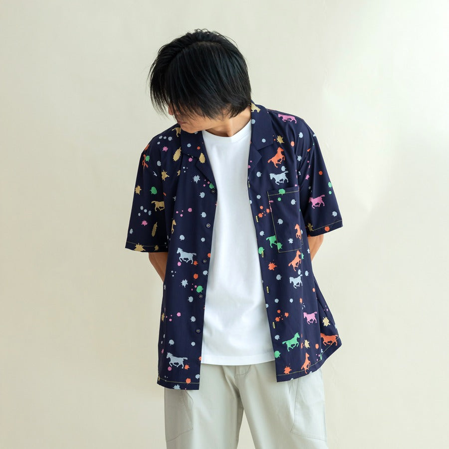 A man wears the men’s Rainbow Horse short-sleeved shirt layered over a white tee, hands in pockets as he looks down with a relaxed expression. The bold neon horse motifs and scattered stars stand out against the navy base, highlighting the shirt’s fun, ca