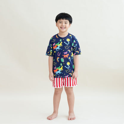 A young boy wearing a navy blue T-shirt with colourful horse and lucky symbol prints paired with red-and-white vertical striped shorts from the Le Petit Society x Sheila Sim CNY 2026 collection stands smiling against a soft cream background.