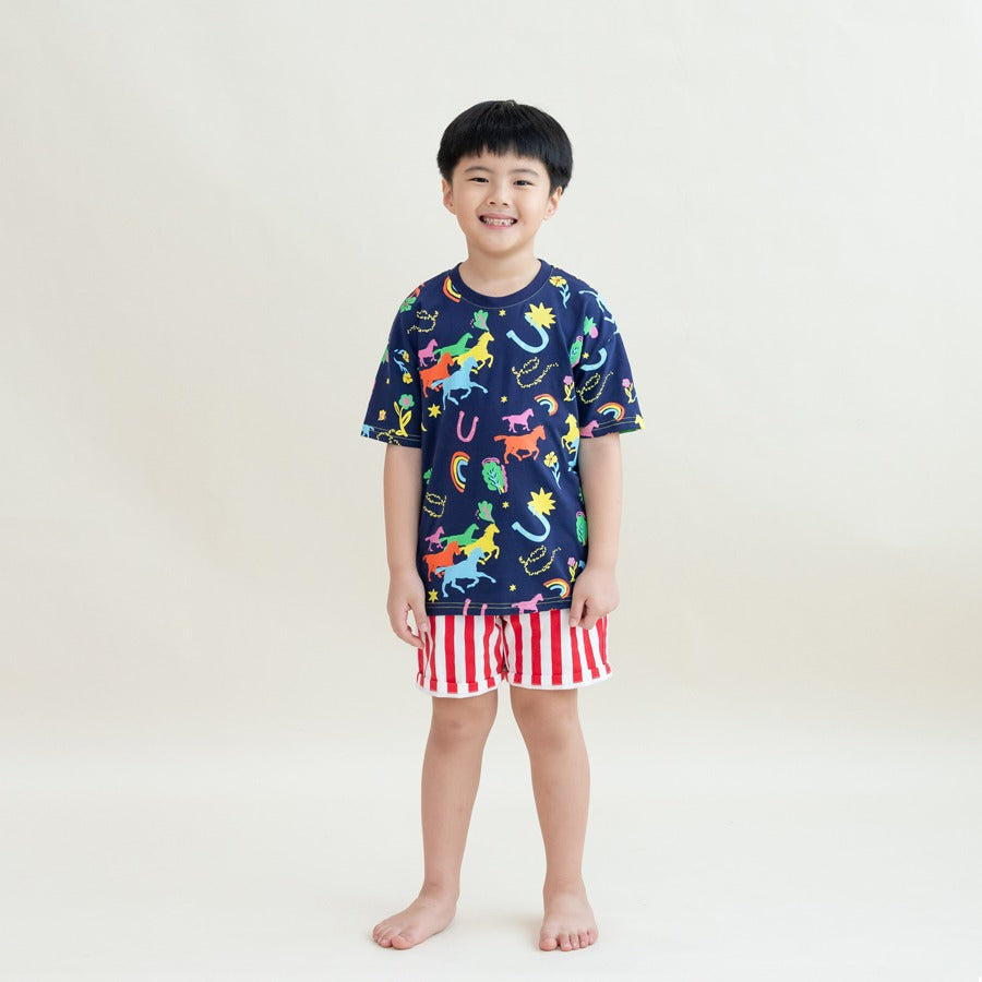 A young boy stands smiling in the Rainbow Horse tee paired with bold red-and-white striped shorts. The vibrant illustrations of horses, stars and rainbows pop against the navy base, giving the outfit a festive, playful look perfect for Lunar New Year.