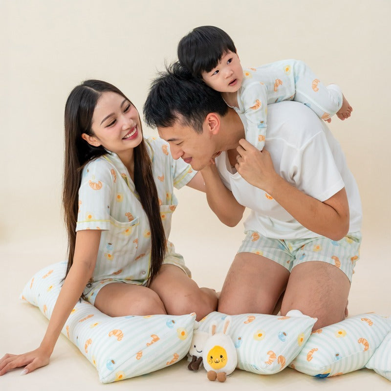 A family of three — mother, father, and baby boy — dressed in matching sleepwear and surrounded by pillows, all featuring a playful breakfast food print from Le Petit Society.