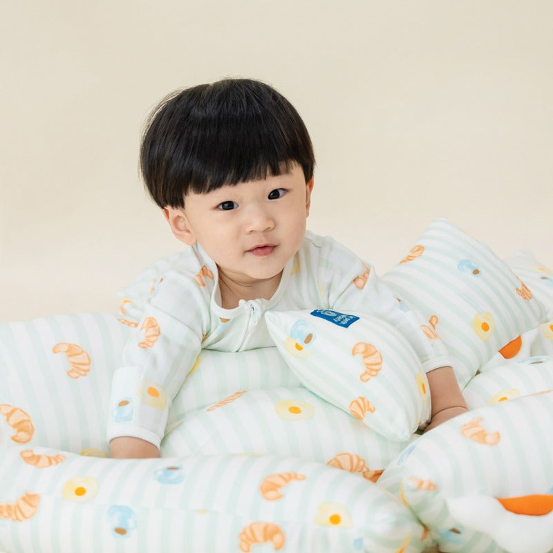 A baby boy in a sleepsuit and surrounded by pillows with a matching print of breakfast food.