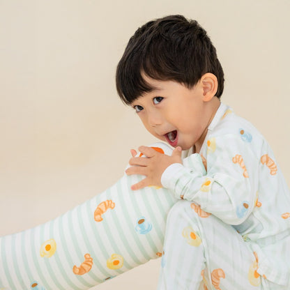A little boy wearing a pyjama set and holding a matching pillow from Le Petit Society. These pieces feature pastel green stripes and a print of breakfast foo.