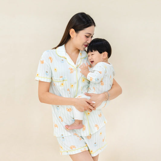 A mother and her baby boy wearing matching sleepwear sets from Le Petit Society, featuring a print of breakfast food and pastel green stripes.