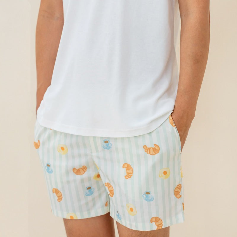 An adult male wearing a pair of men's lounge shorts from Le Petit Society, featuring a print of breakfast food and pastel green stripes.