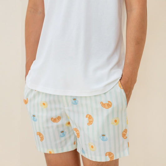 An adult male wearing a pair of men's lounge shorts from Le Petit Society, featuring a print of breakfast food and pastel green stripes.