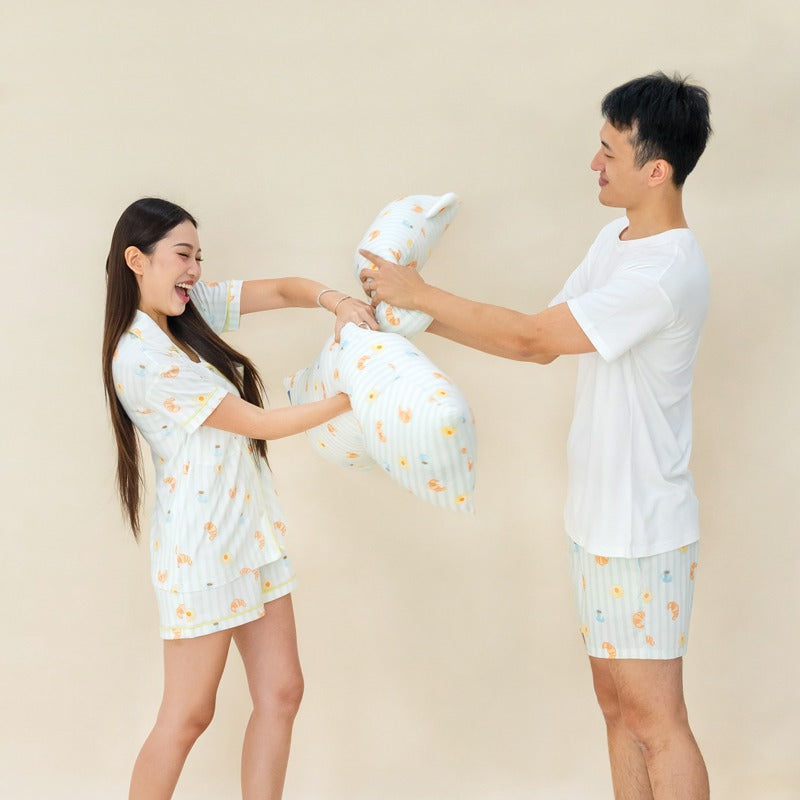 A man and woman having a playful pillow fight, both dressed in matching sleepwear from Le Petit Society featuring a breakfast food print, with pillows in the same design.