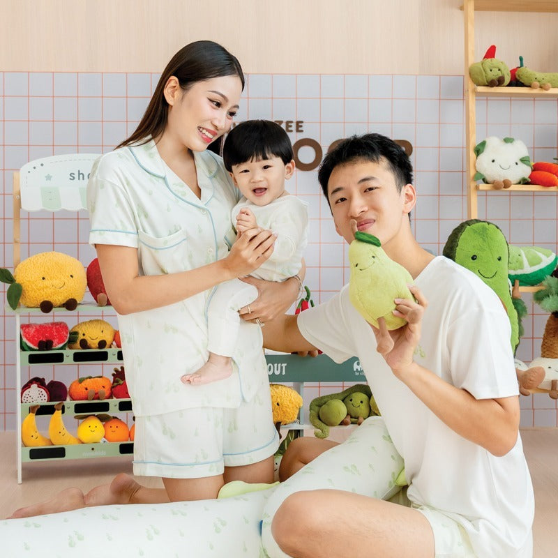 A family of three — mother, father, and baby boy — smiling together while wearing matching outfits from Le Petit Society's sleepwear collection, featuring a print of green pears.