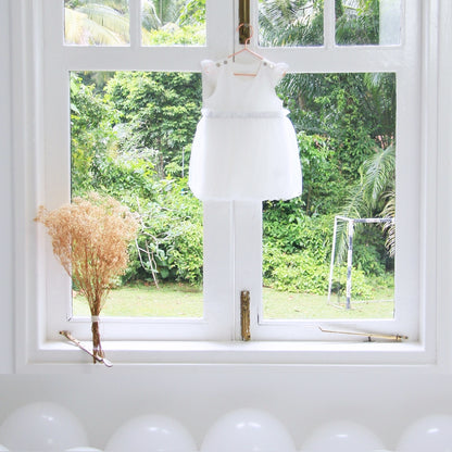 A white baby bubble dress with a tulle skirt and a pair of flutter sleeves is hung on a white window sill of a wedding venue overlooking lush greenery.
