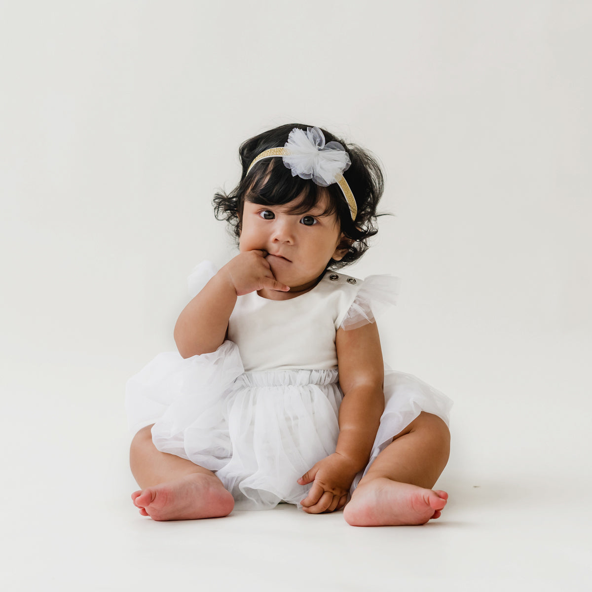 A baby girl looks adorable as she wears a white bubble dress and matching bow headband in white tulle by Le Petit Society.