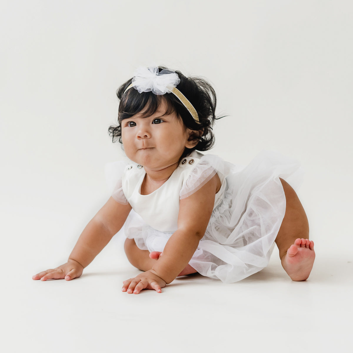 A baby girl looks adorable as she wears a white bubble dress and matching bow headband in white tulle by Le Petit Society.