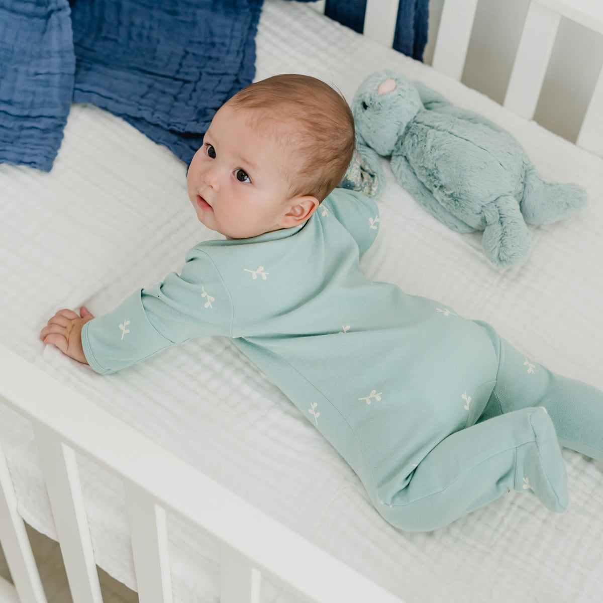 A baby wearing a mint blue organic zip sleepsuit in leaf print from Le Petit Society.