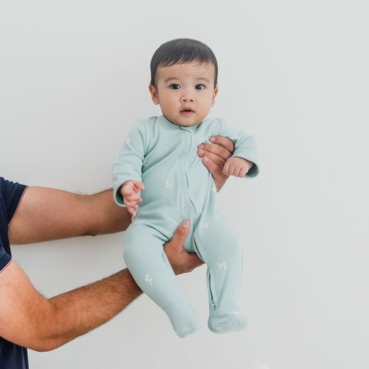 A baby wearing a mint blue organic zip sleepsuit in leaf print from Le Petit Society.